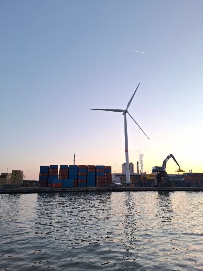 Wind turbine and shipping containers by waterfront at dusk, capturing an industrial scene.